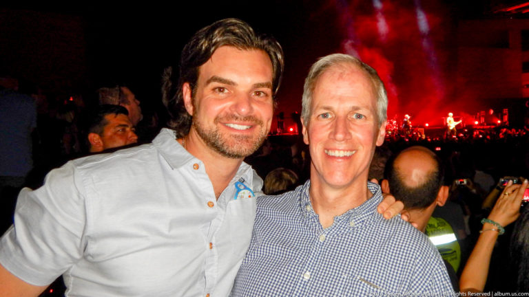 Two smiling men pose in a crowded concert audience with a red-lit stage behind them.