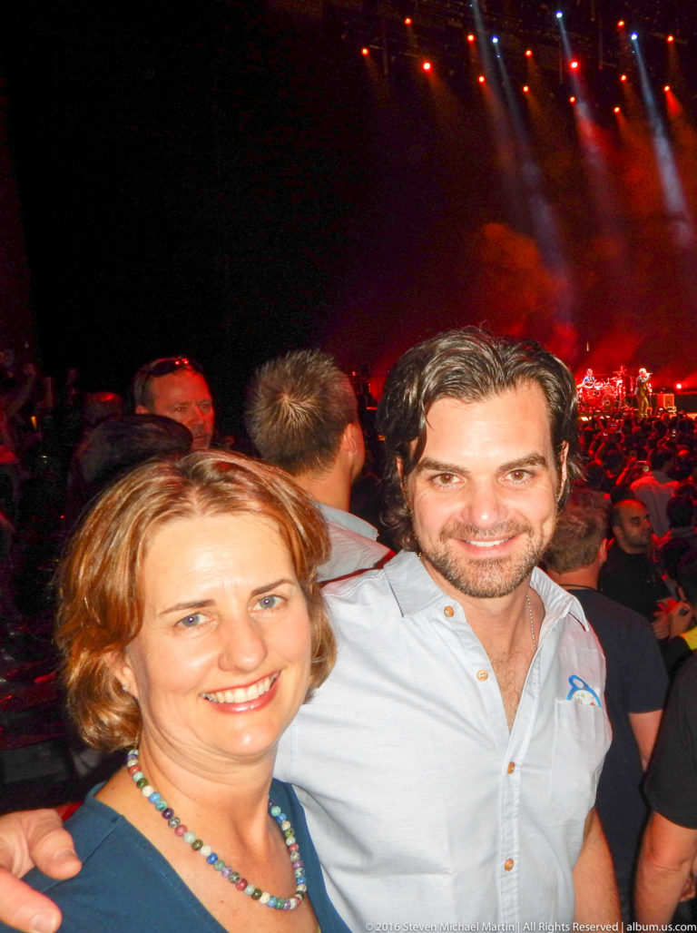 Woman and man smile together in a crowded concert audience with a red-lit stage behind them.
