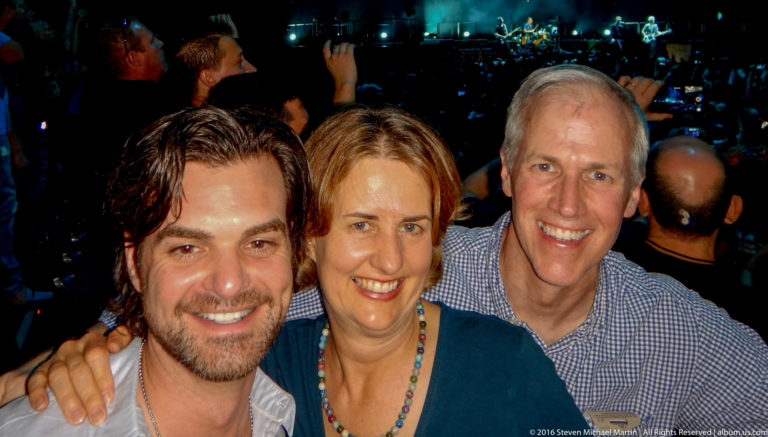 Three smiling adults pose closely together in a concert audience with the stage visible behind them.