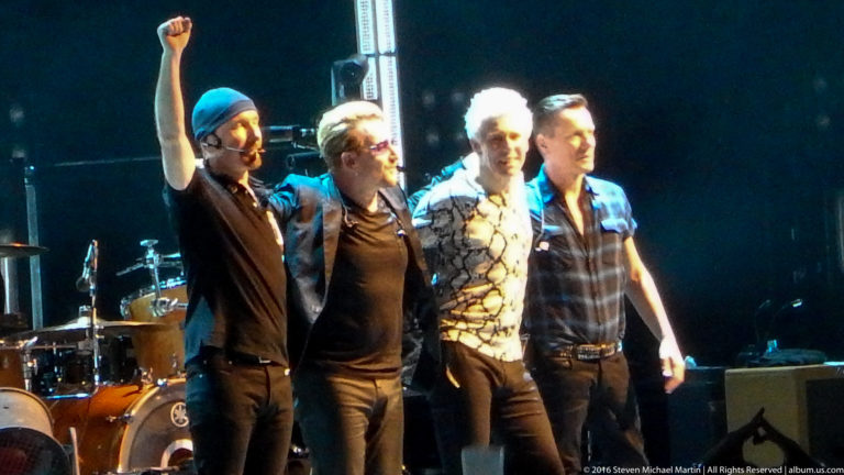 Four male musicians stand together onstage near a drum kit, with one raising his arm.