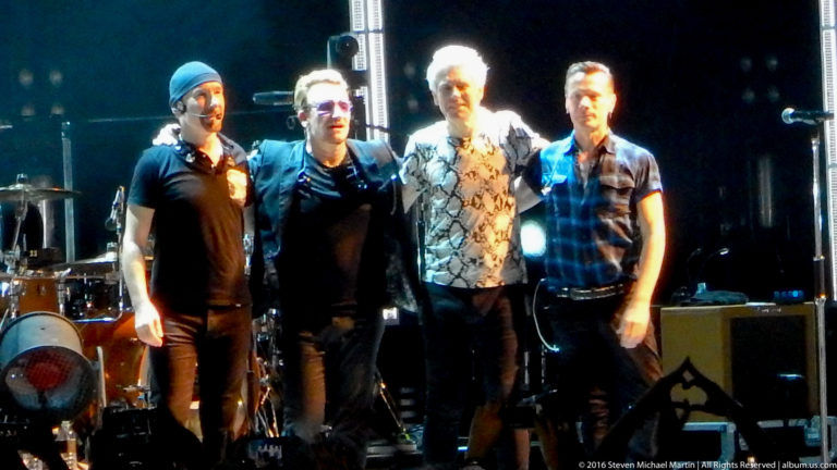 Four male musicians pose together at center stage with drums and amplifiers behind them.