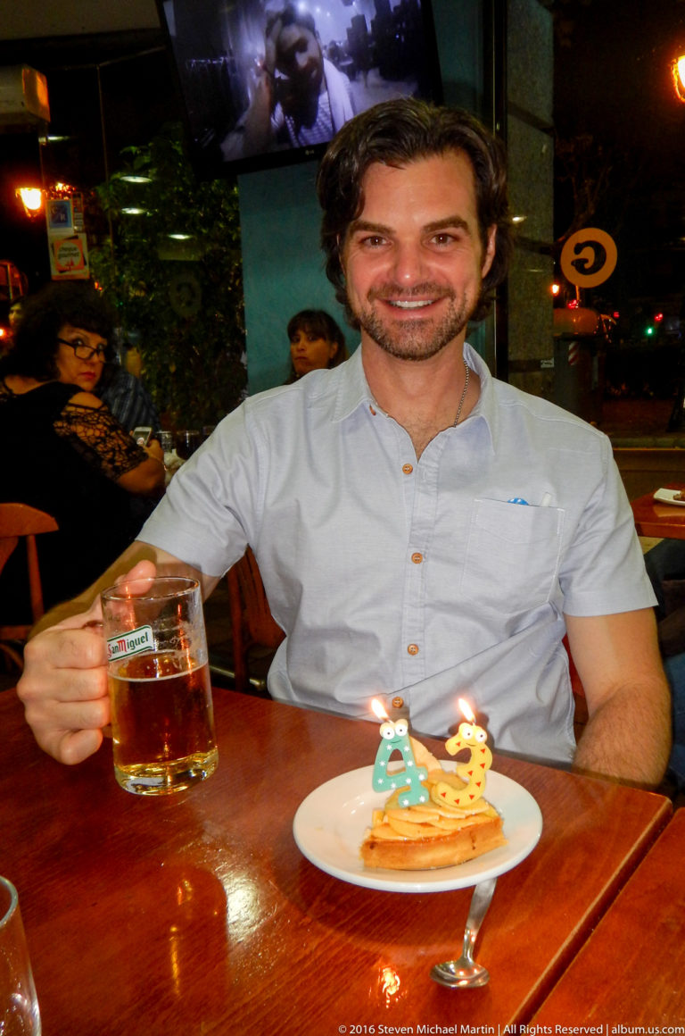 Smiling man holds a beer beside a dessert topped with number candles reading 43.