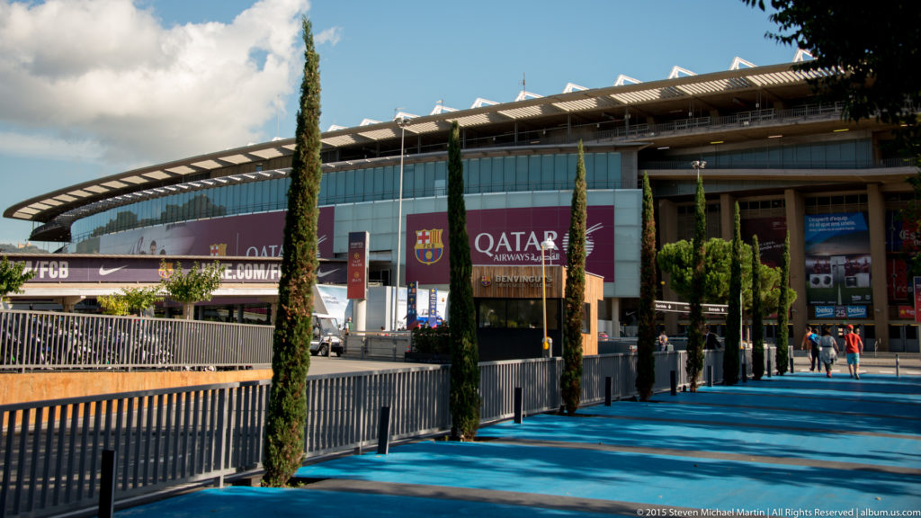 Camp Nou entrance approach with blue walkway, cypress trees, banners, and stadium façade.