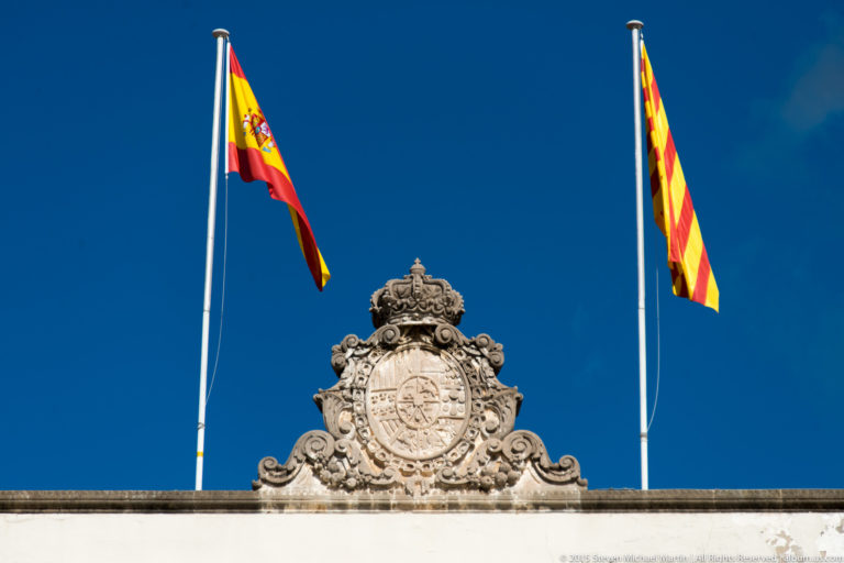 Carved royal crest between Spanish and Catalan flags above a palace roofline.
