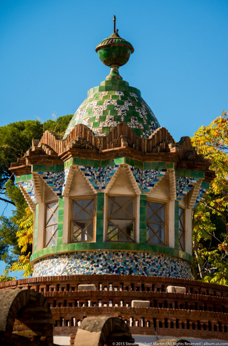 Green, white, and blue mosaic dome above brick arches at the Güell Pavilions.