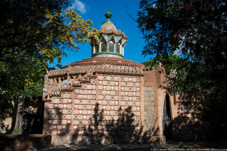 Brick pavilion with circular ceramic decoration and a tiled lantern roof at the Güell Pavilions.