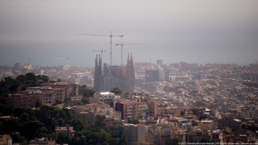 Sagrada Família and construction cranes rising above a hazy Barcelona cityscape.