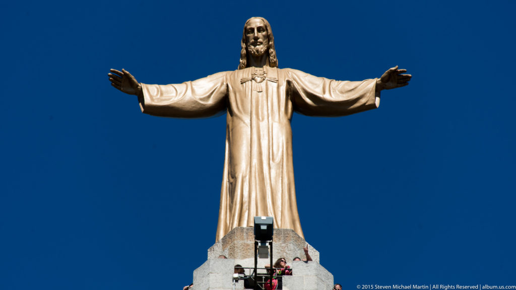 Close view of the Sacred Heart statue of Christ with arms extended against a blue sky.