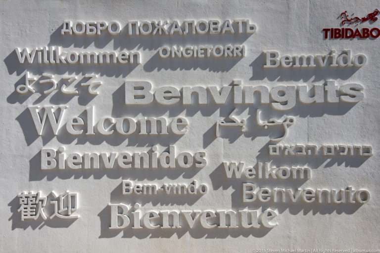Raised multilingual welcome greetings in many scripts on a gray Tibidabo wall.
