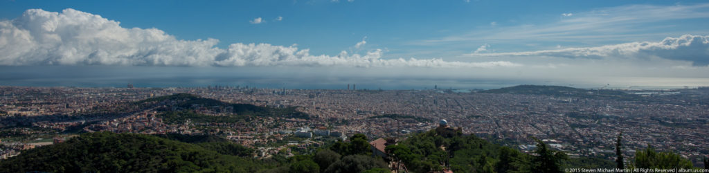 Wide Barcelona panorama from Tibidabo with wooded hills in the foreground and city stretching to the coast.