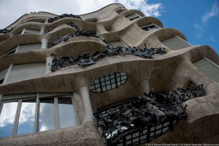 Low-angle view of Casa Milà balconies with tangled dark wrought-iron railings and curved stonework.
