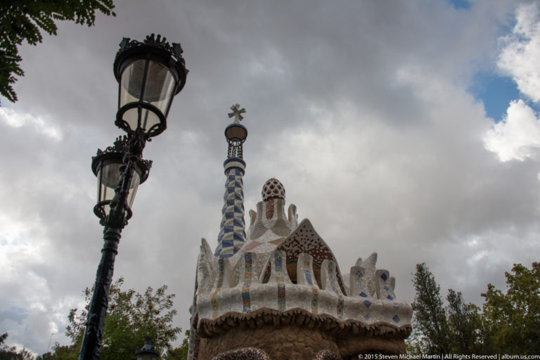 Park Güell entrance pavilion with mosaic roofline and tall blue-and-white tiled chimney beneath cloudy sky.