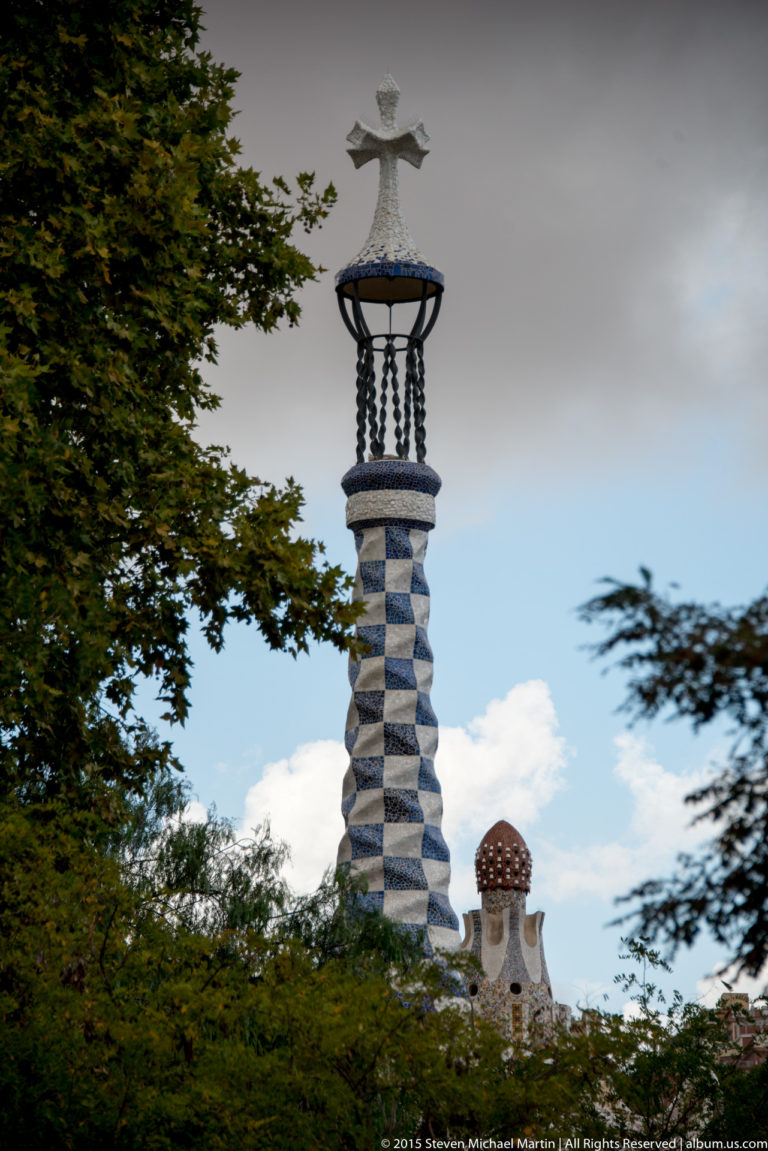 Tall blue-and-white mosaic tower with openwork top rising above trees at Park Güell.