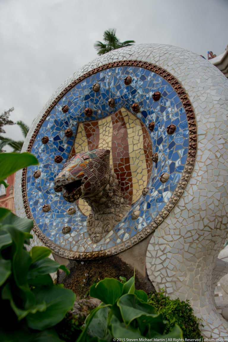 Blue-bordered mosaic roundel with a small projecting animal-head spout on a pale tiled wall.
