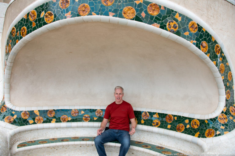 Man in a red shirt seated on a curved white and green mosaic bench at Park Güell.