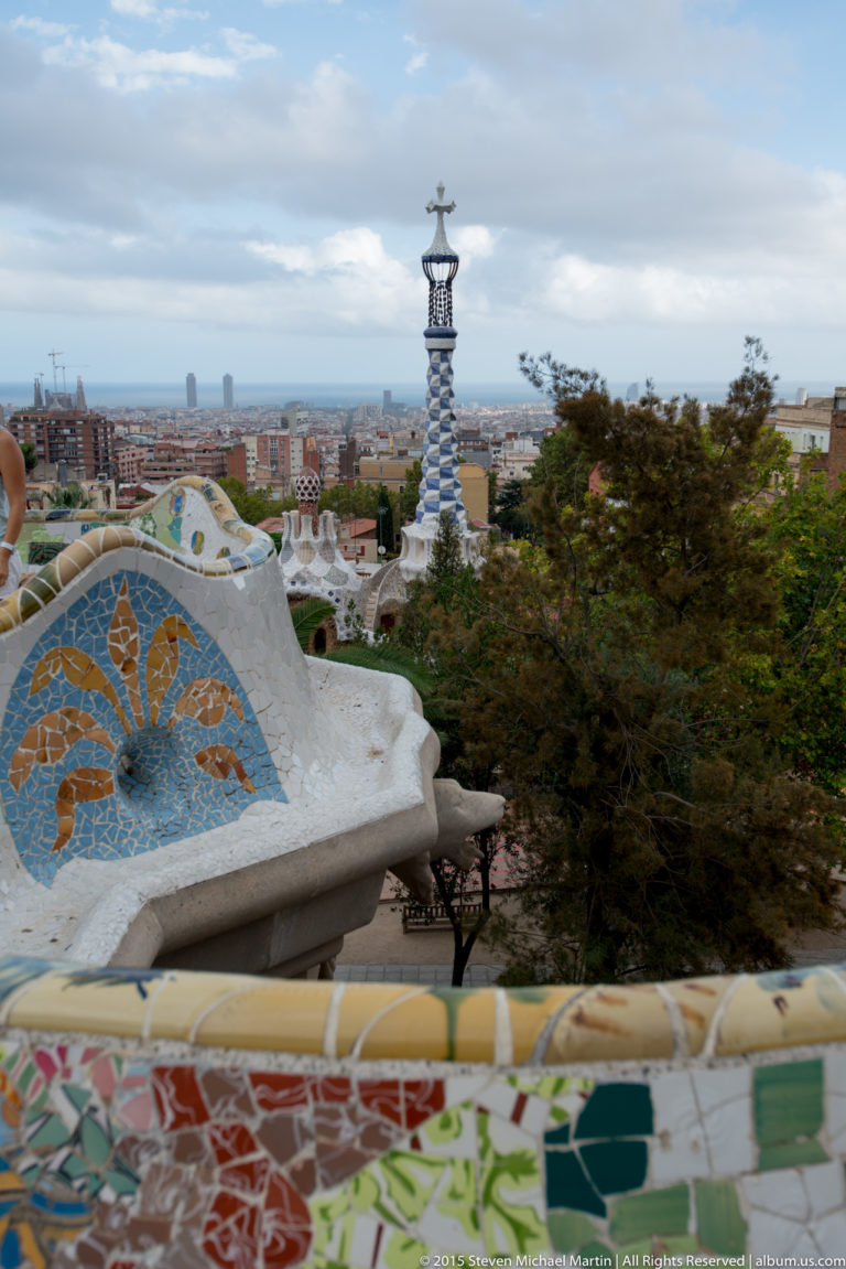 Colorful mosaic terrace edge at Park Güell overlooking Barcelona with a tiled tower beyond.