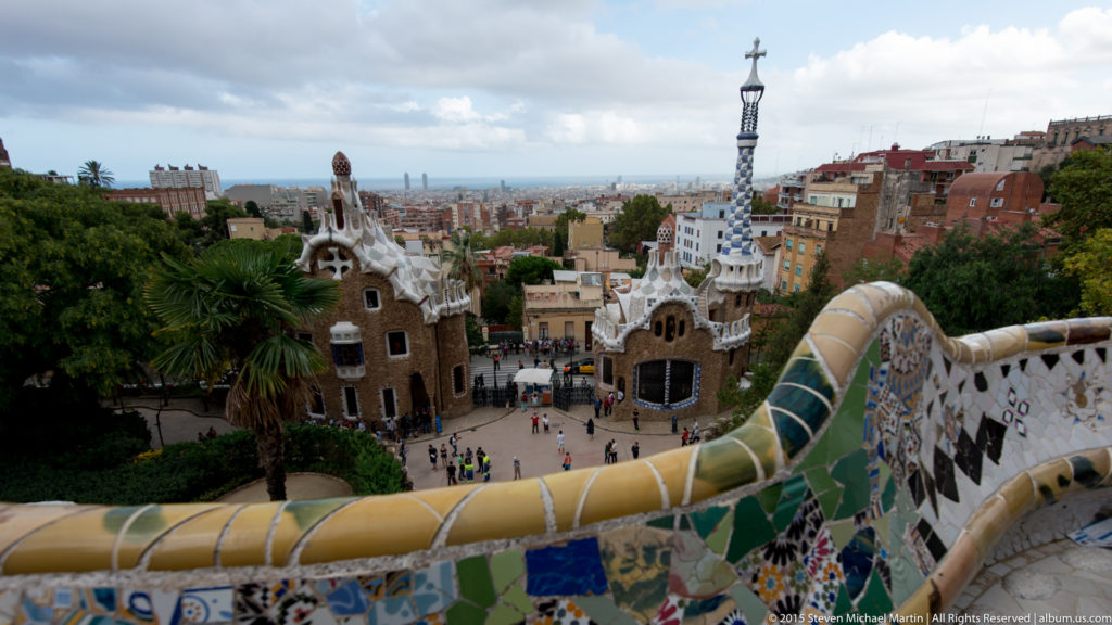 Park Güell entrance pavilions below a mosaic terrace rail with Barcelona spreading into the distance.