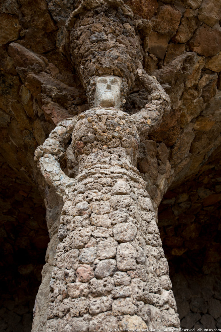 Rough stone column shaped like a standing figure beneath a Park Güell viaduct.