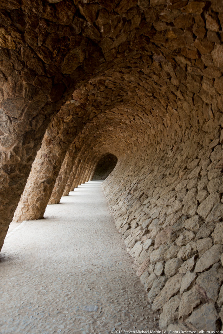 Curving covered stone passage with leaning columns beneath a Park Güell viaduct.