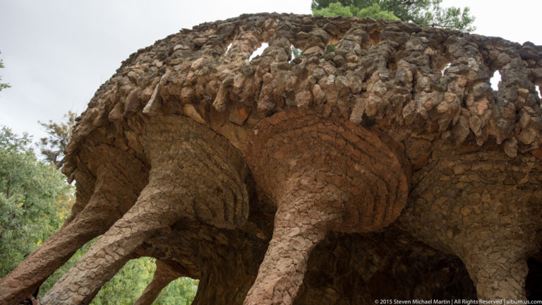 Branching rough-stone columns support a rocky viaduct structure amid greenery at Park Güell.