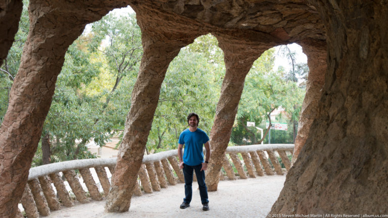 Man standing beneath large leaning rough-stone columns along a covered Park Güell passage.