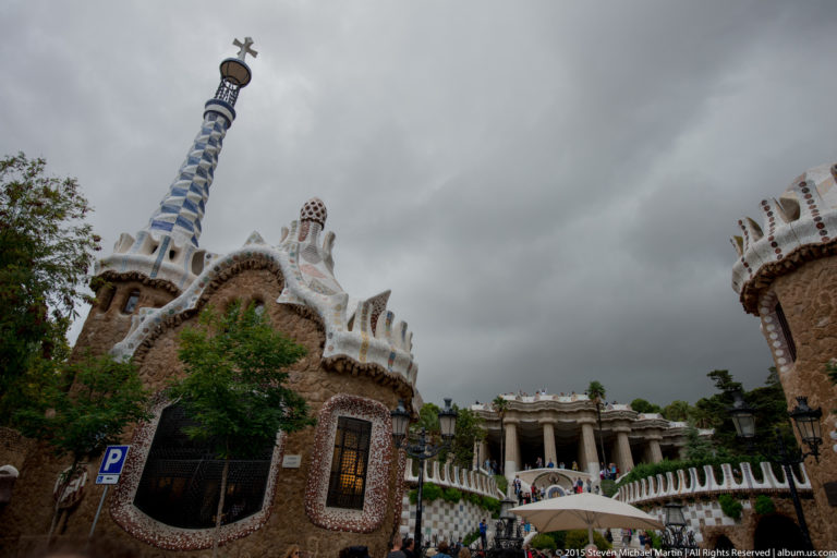 Park Güell entrance pavilion, mosaic roofline, and Hypostyle Room beneath a cloudy sky.