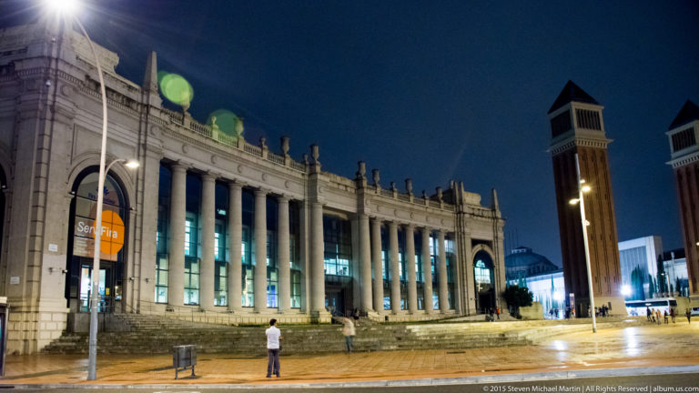 Illuminated colonnaded exhibition hall beside a Venetian Tower on Montjuïc at night.
