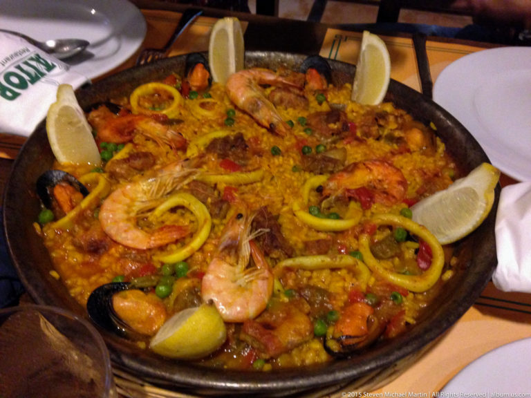 Pan of seafood paella with prawns, mussels, squid rings, peas, peppers, and lemon wedges.