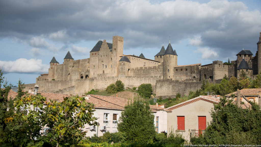Stone walls and towers of Carcassonne rise above white houses and red-tiled roofs under a partly cloudy sky.