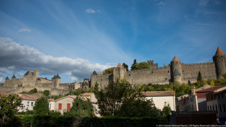 Long stone rampart with conical-roofed towers above houses and trees at Carcassonne beneath a broad blue sky.