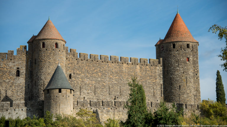 Two round stone towers with red conical roofs flank a crenellated wall, with a smaller turret below.