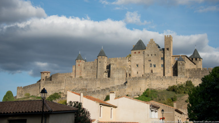 Carcassonne’s dense stone walls and towers rise above low houses beneath dramatic clouds.