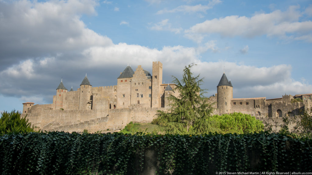 Panoramic view of Carcassonne’s stone ramparts and many roofed towers rising above trees beneath broken clouds.