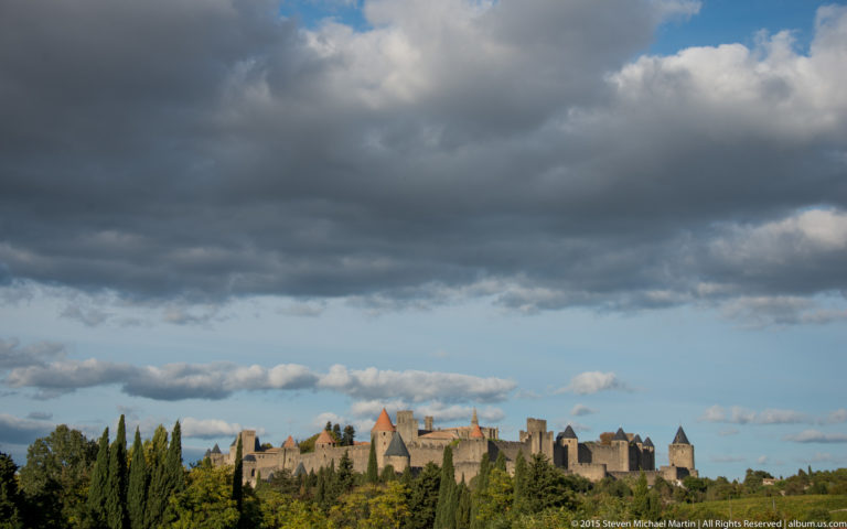 Distant Carcassonne skyline above trees beneath a vast blue sky filled with large dark and white clouds.
