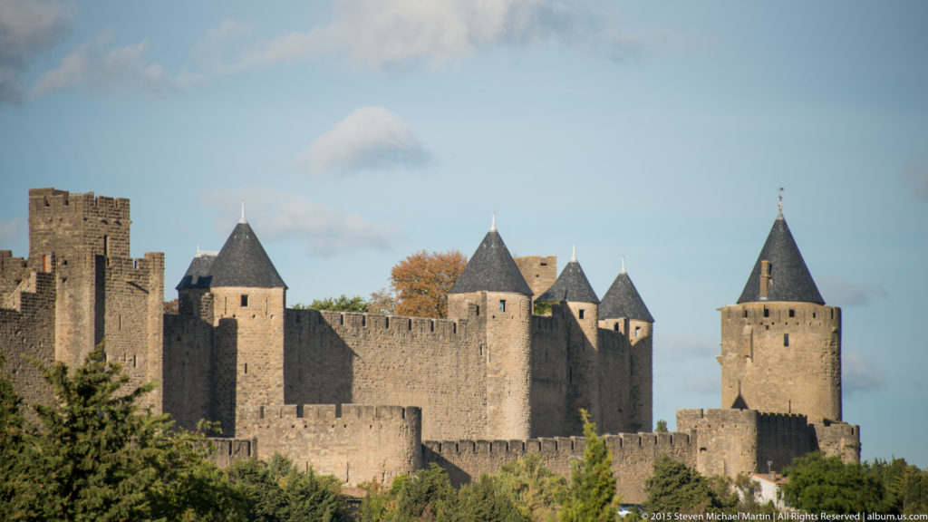 Cluster of Carcassonne round and square towers linked by high stone walls, with trees and autumn foliage below.