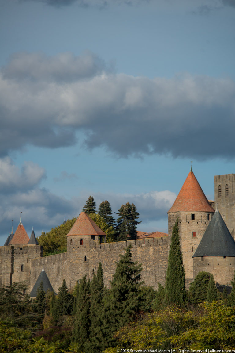 Stone ramparts and red-roofed round towers at Carcassonne rise behind dense trees under a dark cloud band.