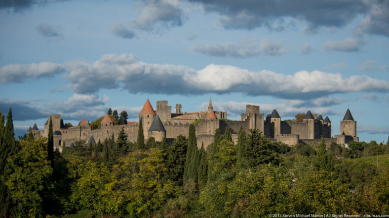 Broad view of Carcassonne’s stone walls and many towers rising above dense trees beneath large white clouds.