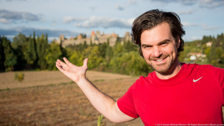 Smiling visitor in red extends an open hand toward the softly focused fortified skyline of Carcassonne behind him.