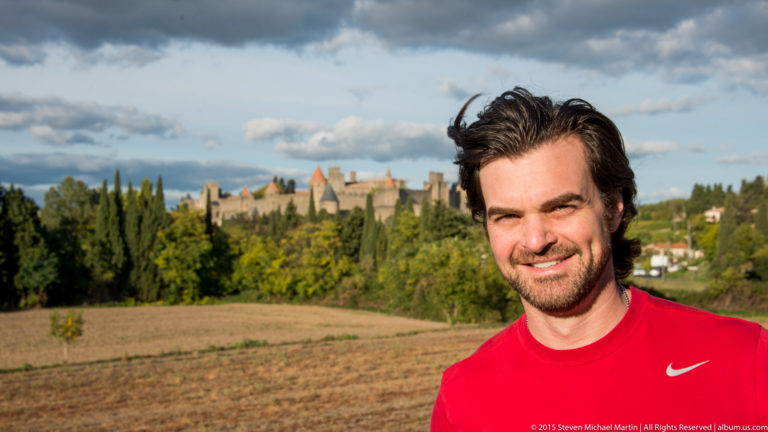 Smiling visitor in a red shirt stands before fields and trees with Carcassonne’s distant towers and walls behind.