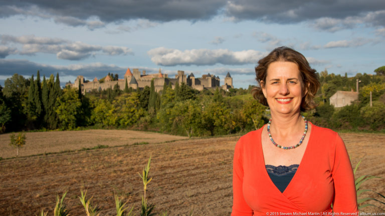 Visitor stands before a cultivated field with the distant fortified skyline of Carcassonne extending above trees.