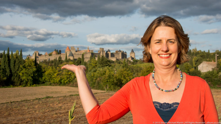 Smiling visitor raises one hand toward the distant towers and walls of Carcassonne across a field and trees.