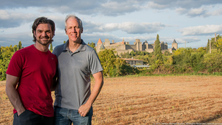 Two visitors stand in an open field with the distant towers and ramparts of Carcassonne visible behind them.