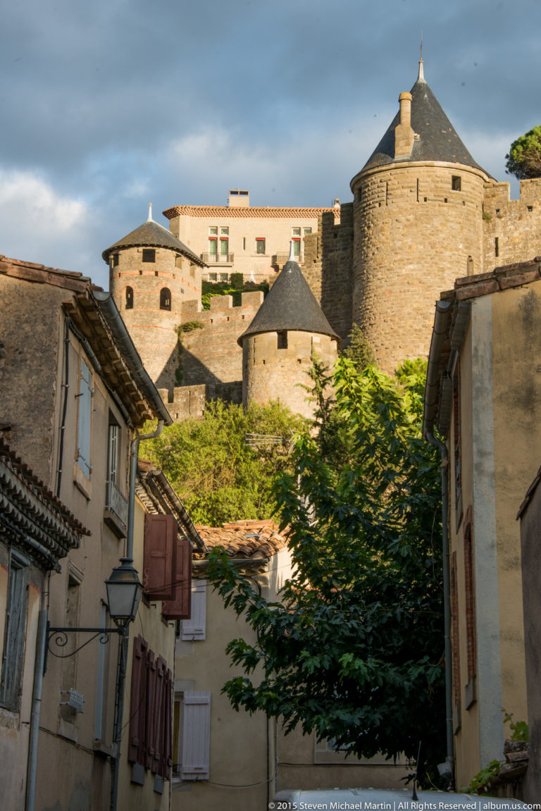 Narrow Carcassonne street with shuttered houses and trees below large round towers and stone fortification walls.