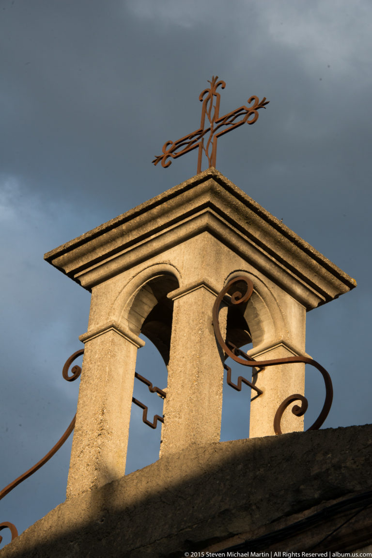 Small twin-arched bell gable with decorative ironwork and a cross-like finial against a dark sky.