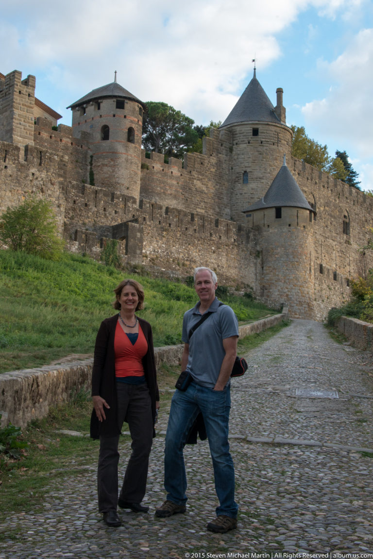 Two visitors stand on a cobbled path below Carcassonne’s stone walls and round defensive towers.