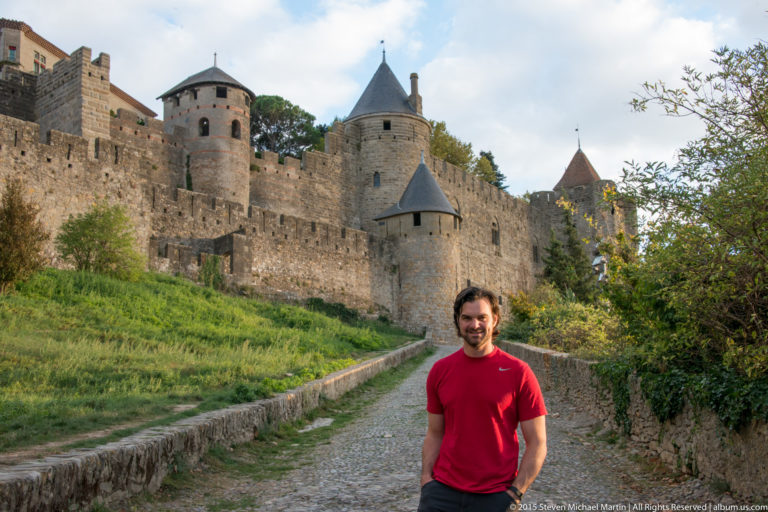 Visitor in red stands on a gravel path below Carcassonne’s high stone walls and conical-roofed towers.