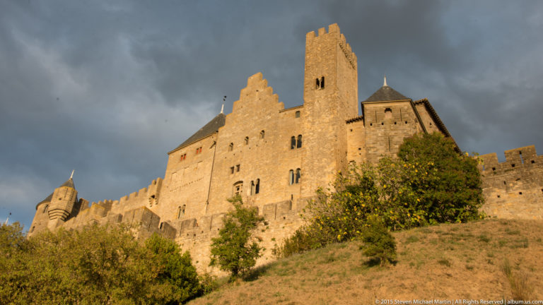 Tall stone building with square tower and stepped wall profile rises above a grassy slope at Carcassonne.