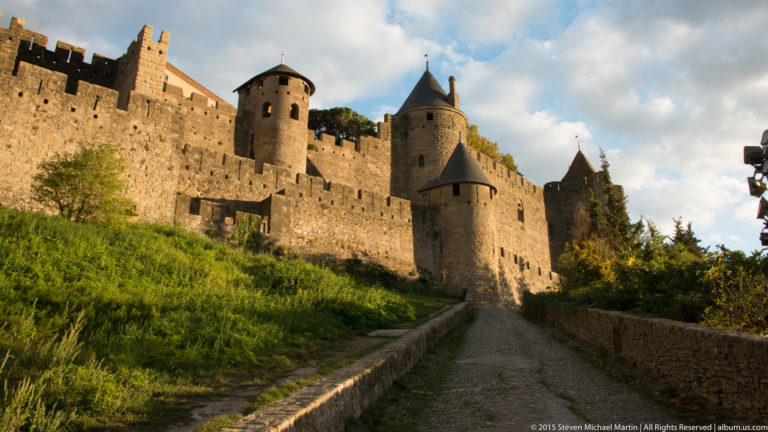 Path rises beside grassy banks toward Carcassonne’s stone walls and several conical-roofed defensive towers.
