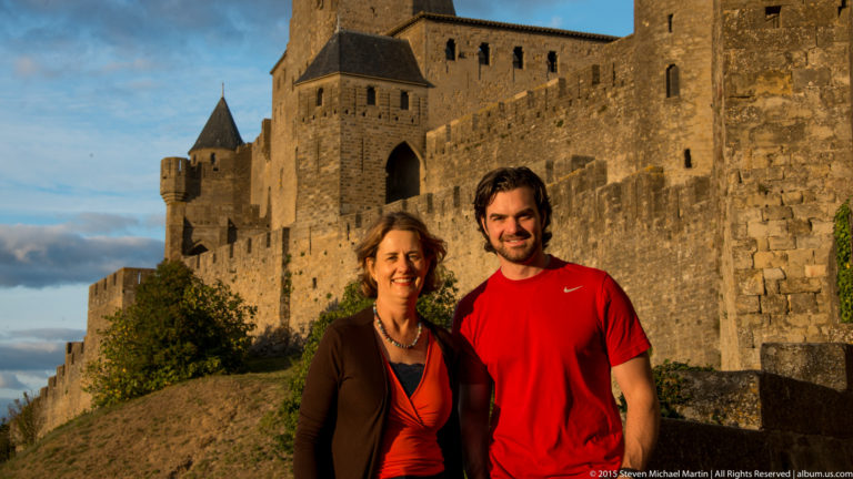 Two visitors stand together below Carcassonne’s high stone walls and towers in warm evening light.
