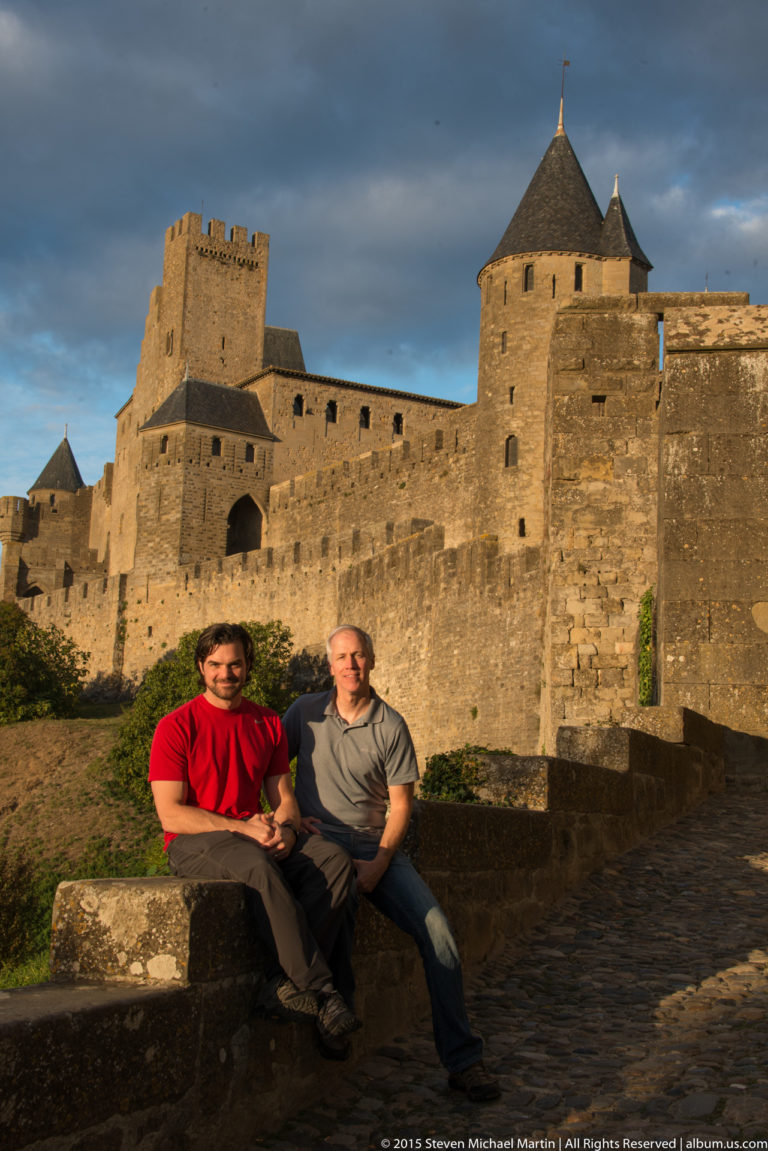 Two visitors sit on a low stone wall before Carcassonne’s long ramparts, square tower, and round roofed tower.
